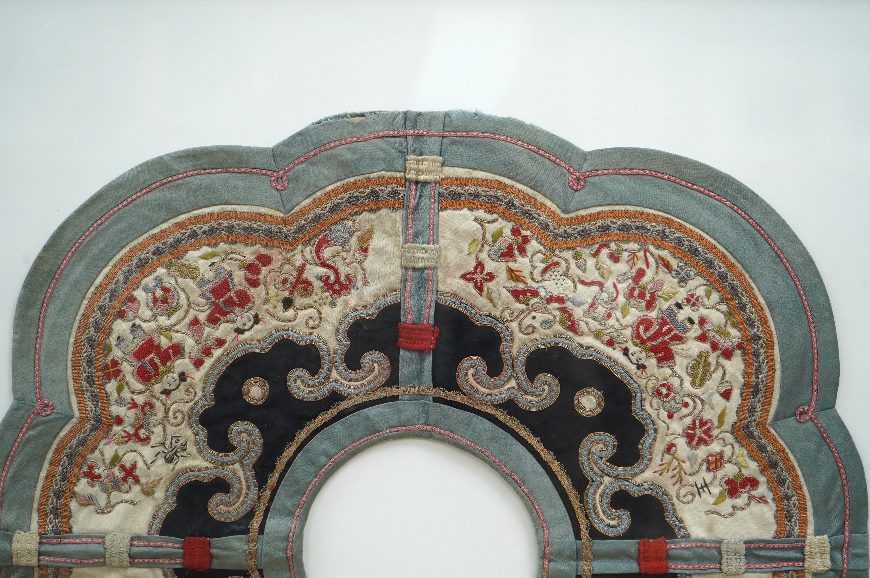 An unusual framed Chinese embroidered cloud shaped child's collar, embroidered with figures and auspicious symbols, in multi-coloured silks and gold thread, edged with braiding and a green silk border, collar 35cm wide.